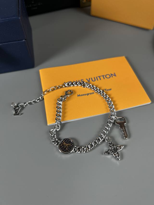 Lv Monogram Lock 手链玩趣汇聚品牌元素，以经典Monogram帆布中的Lv字母和 Monogram 花卉铭纹点明旅行硬箱的设计传承，搭配可调节链